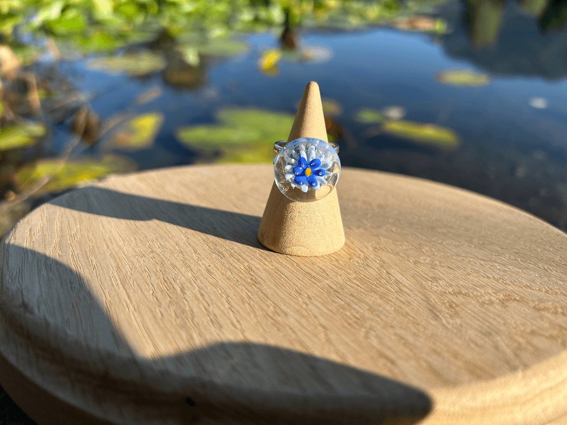 Bague cabochon en verre transparent à motif floral bleu présentée de face sur un cône en bois.