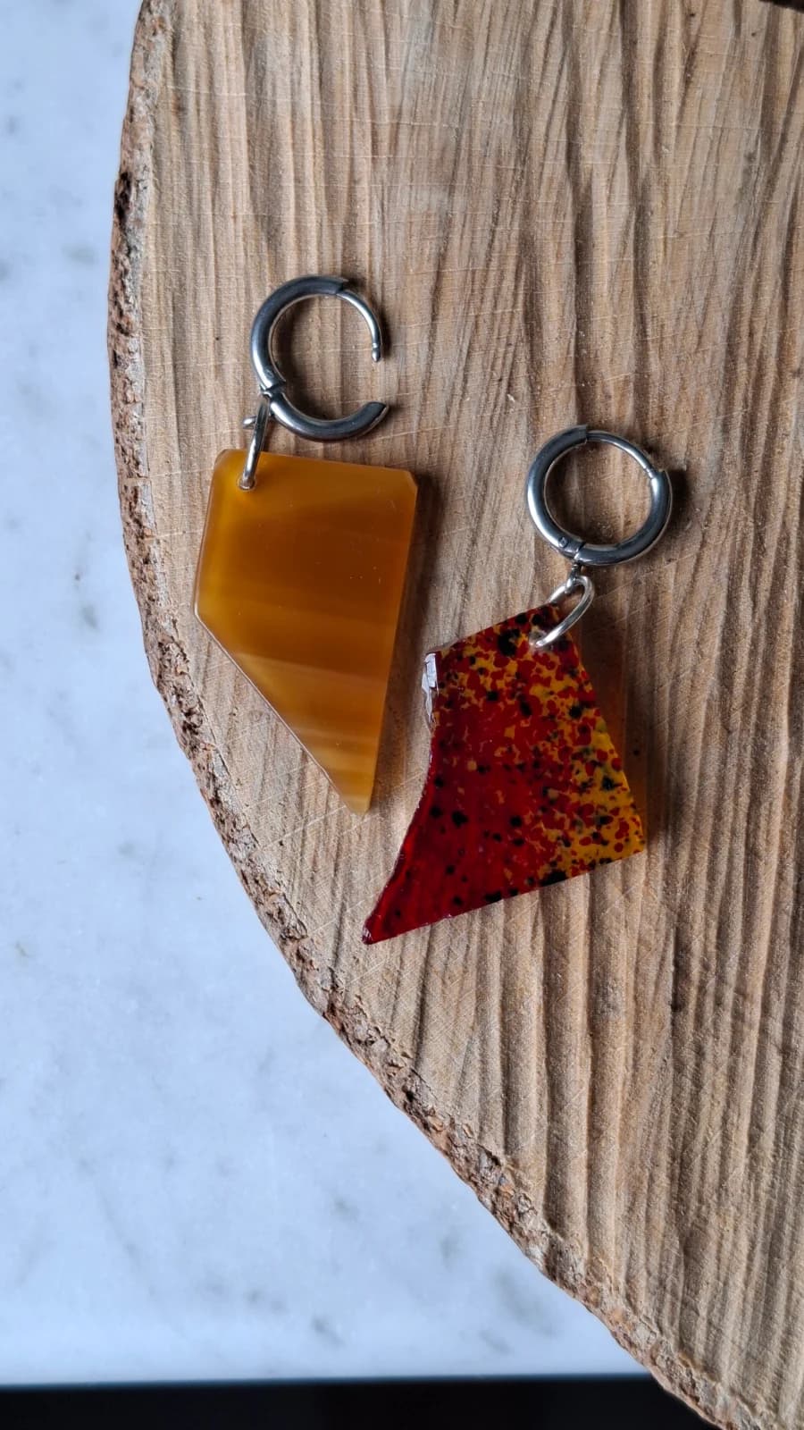 Boucles d'oreilles dormeuses asymétriques en verre géométrique jaune et rouge moucheté posées sur du bois.