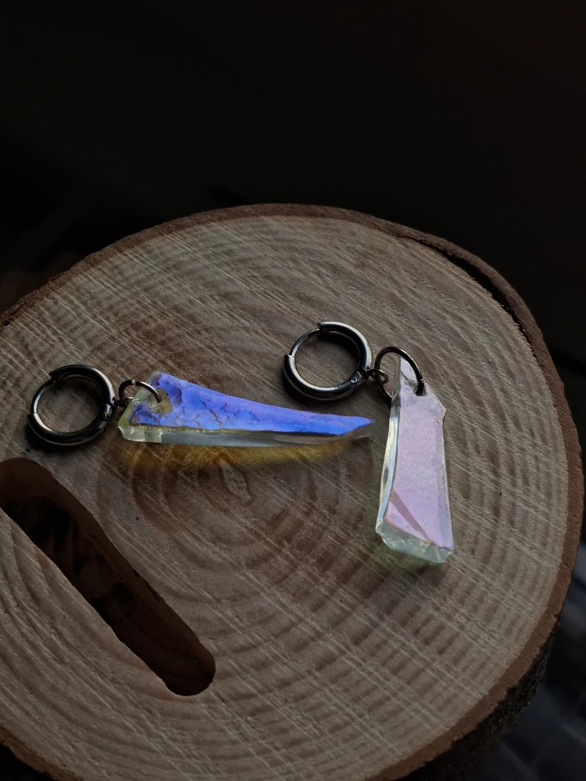 Paire de boucles d'oreilles en verre aux reflets violets et bleus sur socle en bois dans la pénombre.