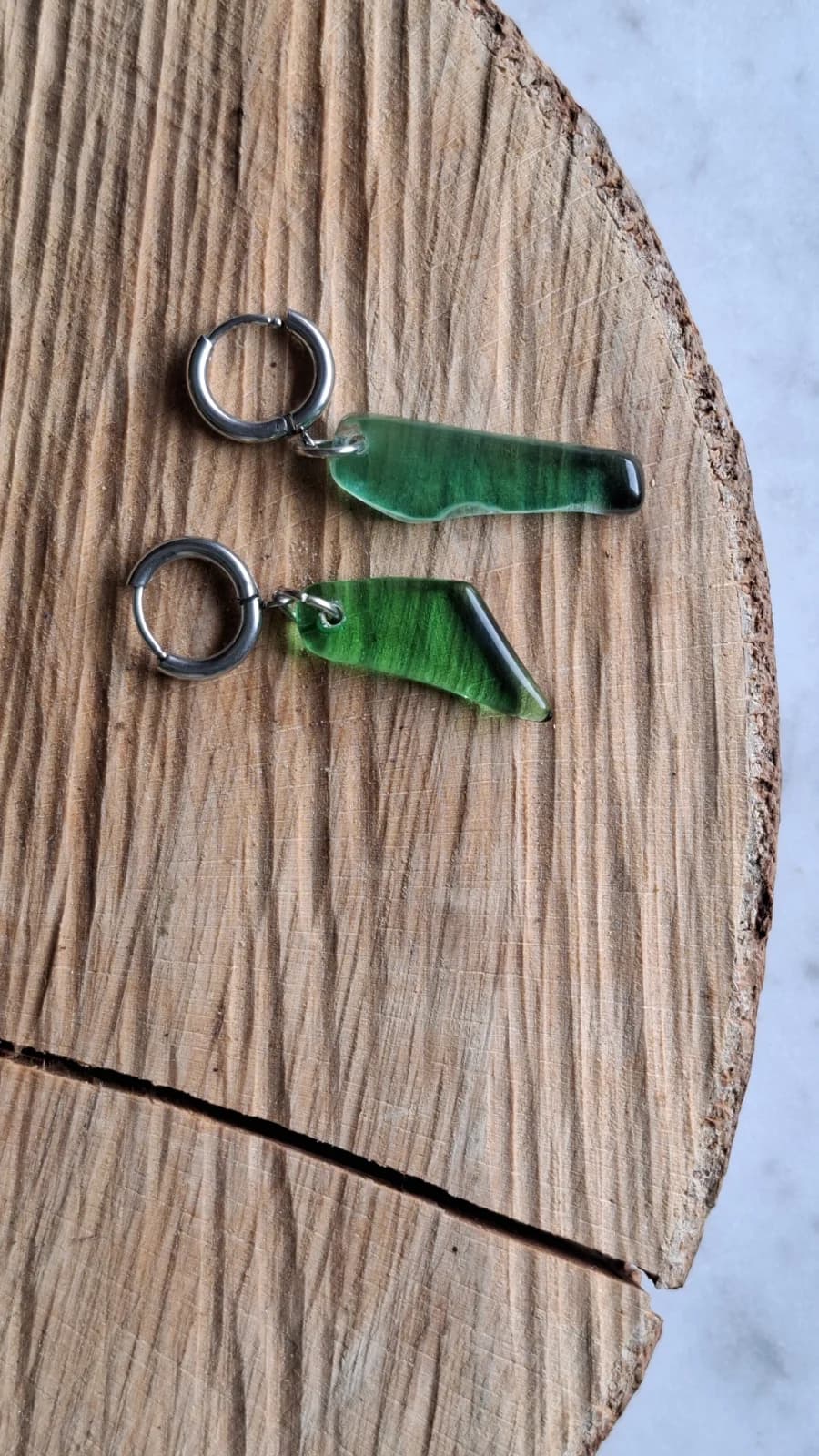 Boucles d'oreilles artisanales en verre vert et acier inoxydable posées sur une rondelle de bois.