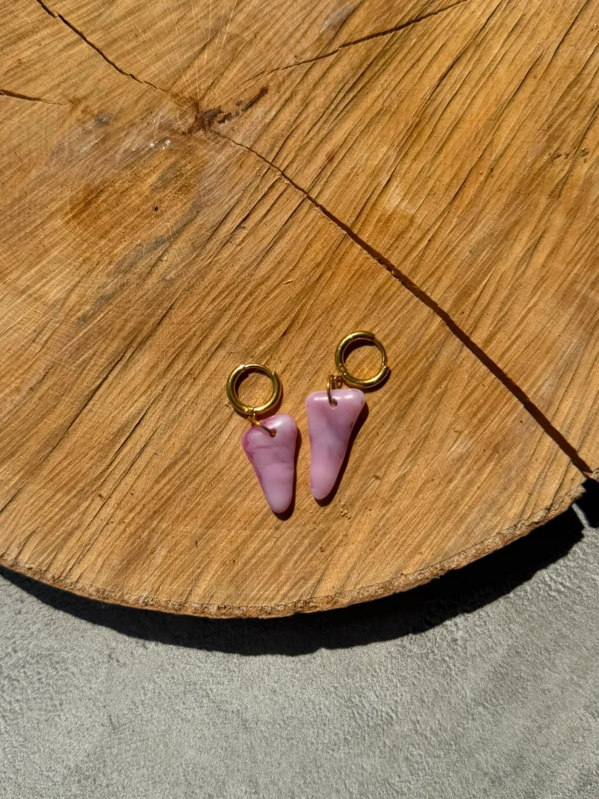 Boucles d'oreilles en verre marbré rose et anneaux dorés posées à plat sur un socle en bois brut.