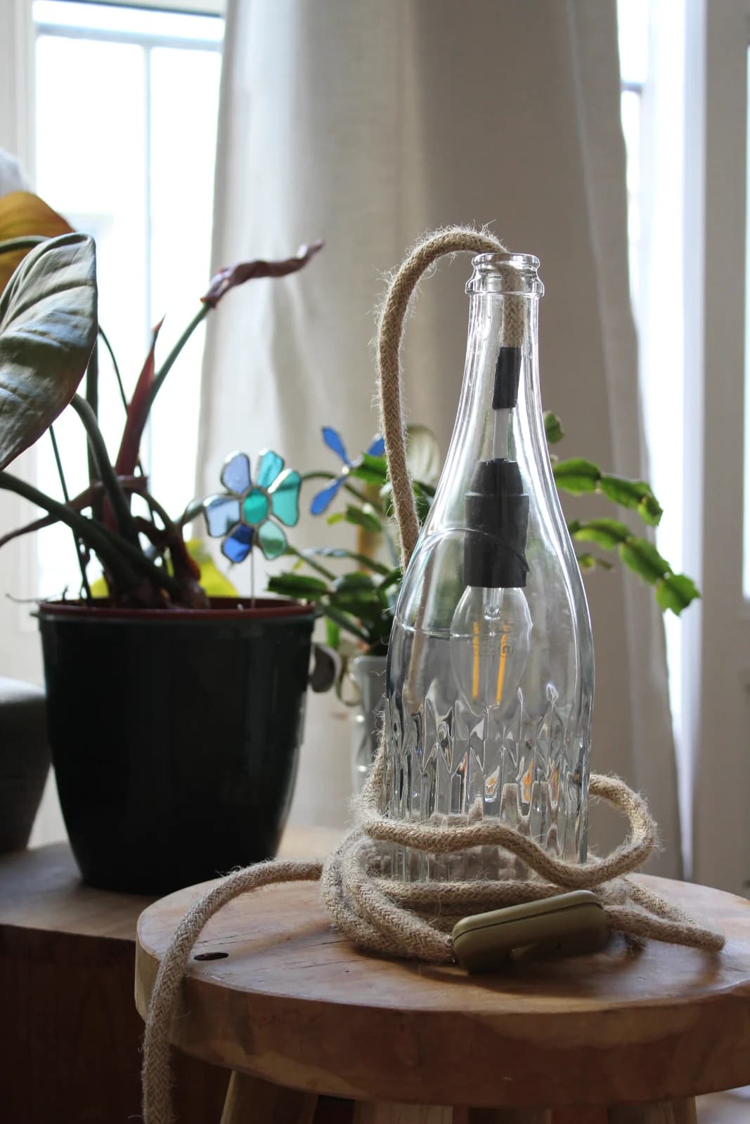 Lampe à poser en bouteille de verre transparent avec câble en corde naturelle posée sur un tabouret en bois.