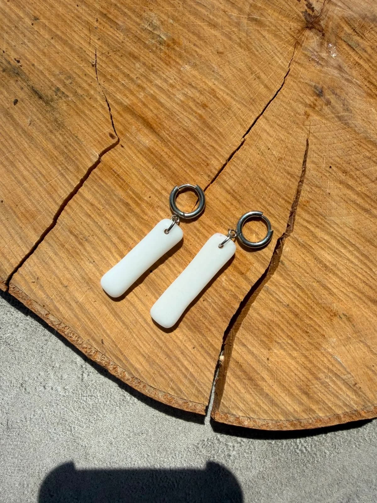 Boucles d'oreilles pendantes blanches en verre montées sur anneaux argentés, posées sur une rondelle de bois.