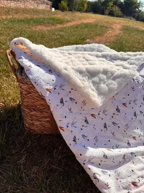 Couverture bébé à motifs sylvestres posée dans un panier en osier en extérieur, avec revers polaire texturé.