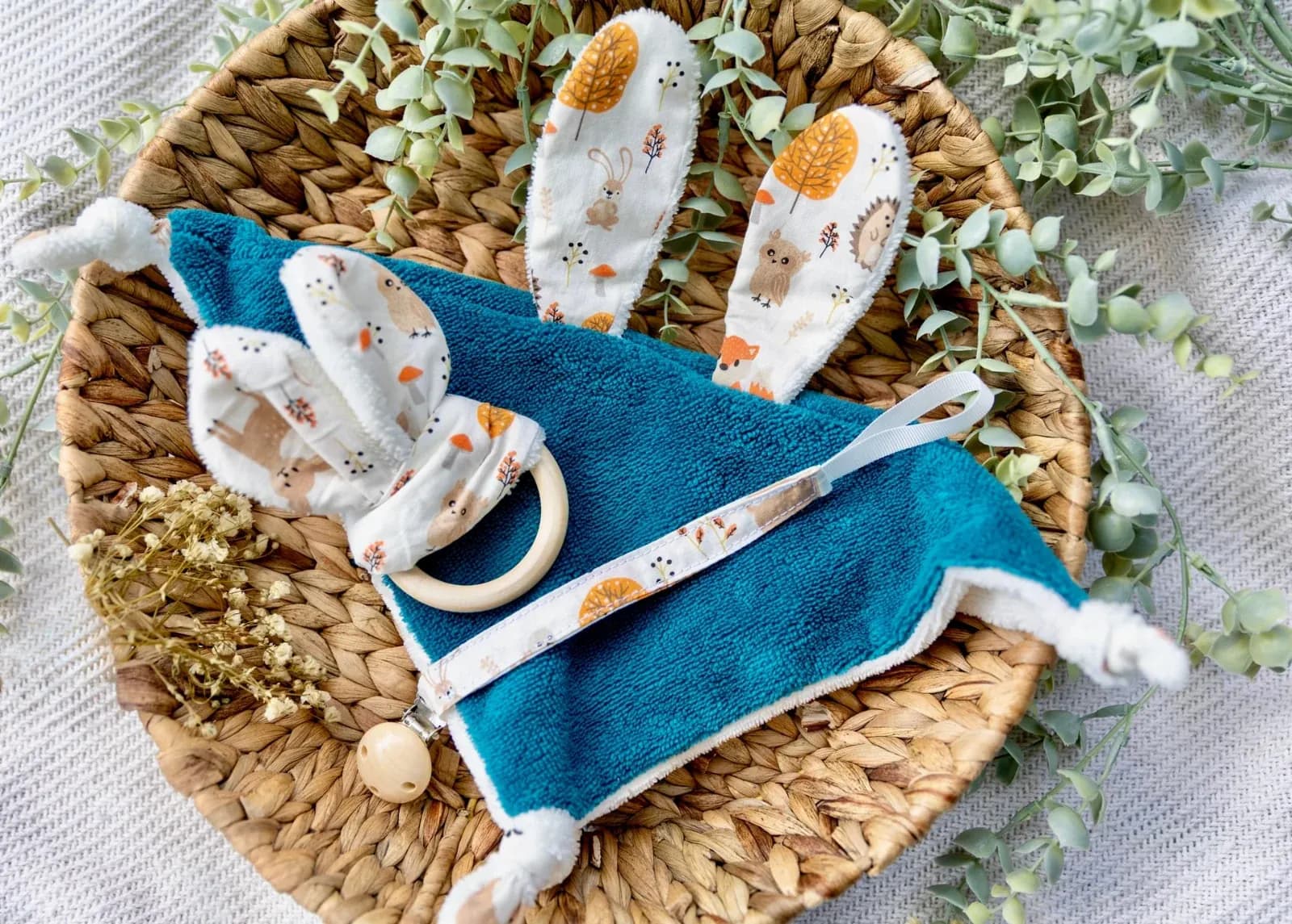 Doudou plat bleu canard à oreilles de lapin, hochet anneau et attache-sucette aux motifs d'automne dans une corbeille.