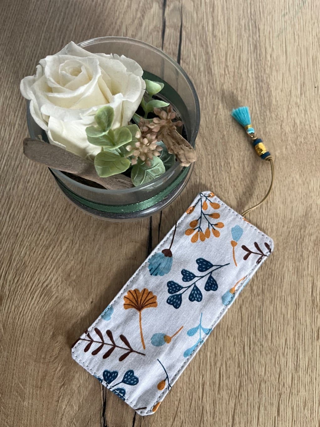 Marque-page en tissu à motifs de feuilles bleues et moutarde avec pompon turquoise posé sur une table en bois.