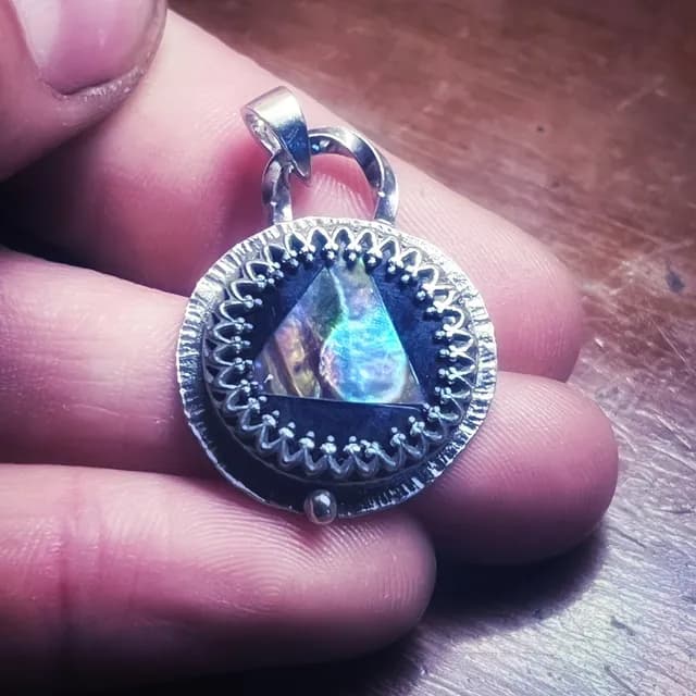 Pendentif médaillon en argent ciselé tenu entre les doigts, révélant les reflets irisés de l'abalone.