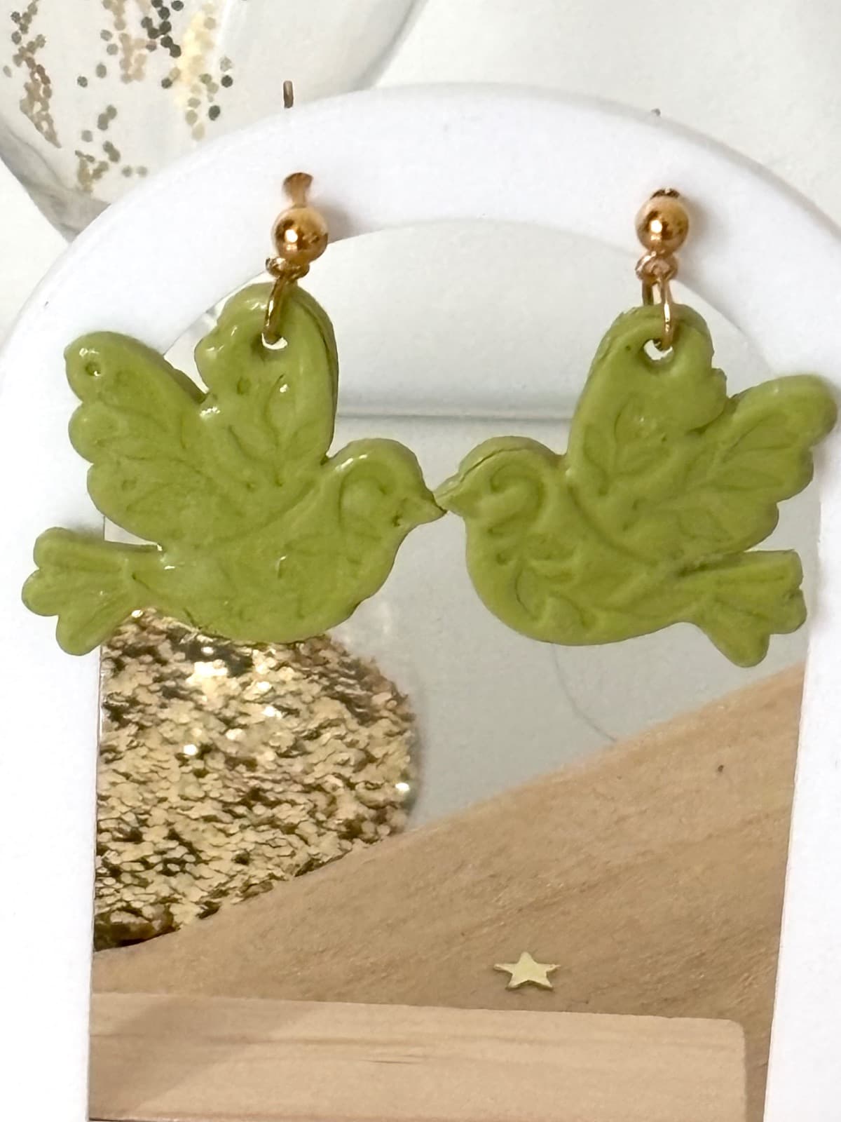 Boucles d’oreilles oiseaux vert olive suspendues face à face sur un support blanc