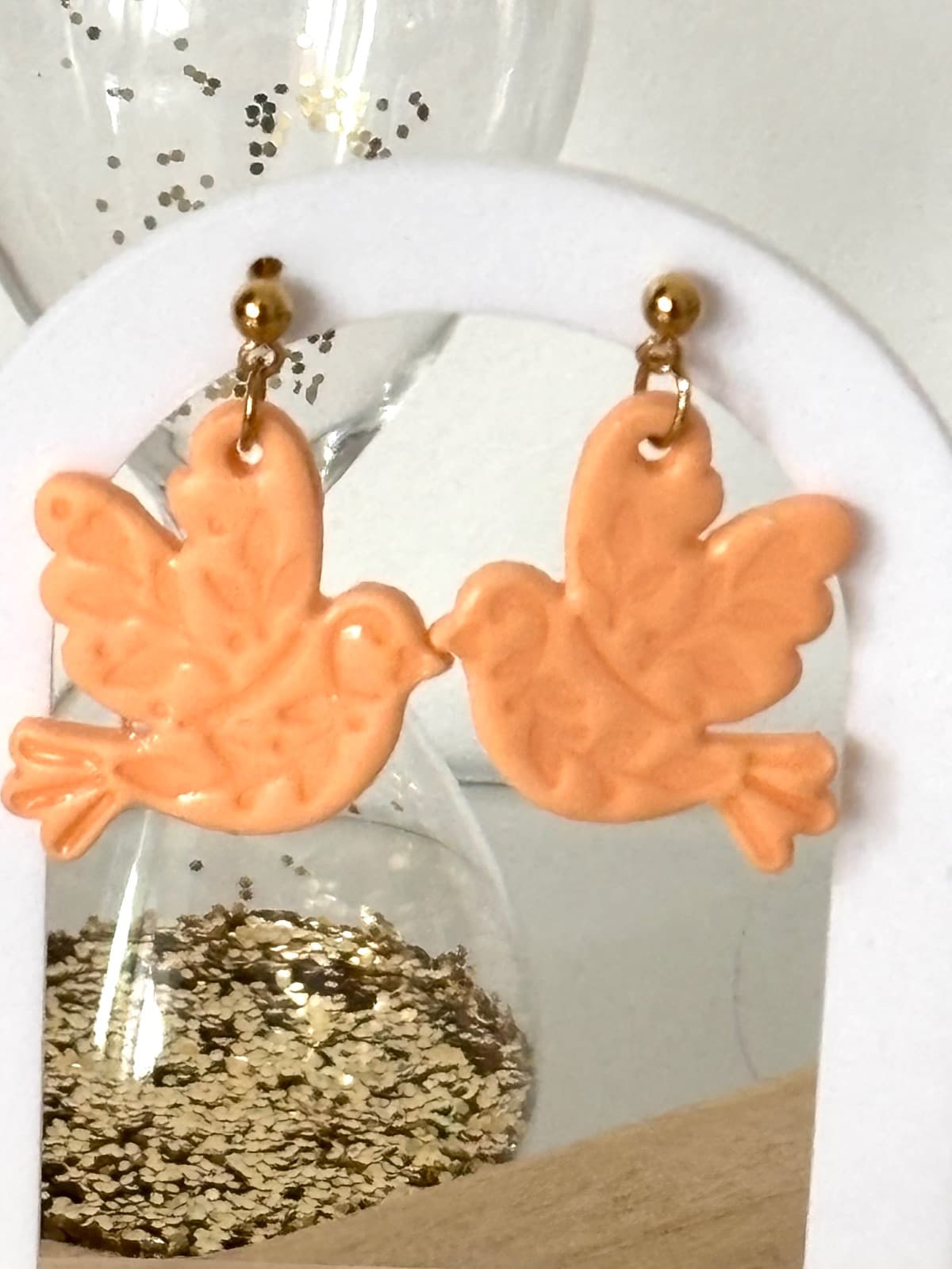 Boucles d'oreilles oiseaux couleur pêche suspendues à un support blanc avec attaches dorées