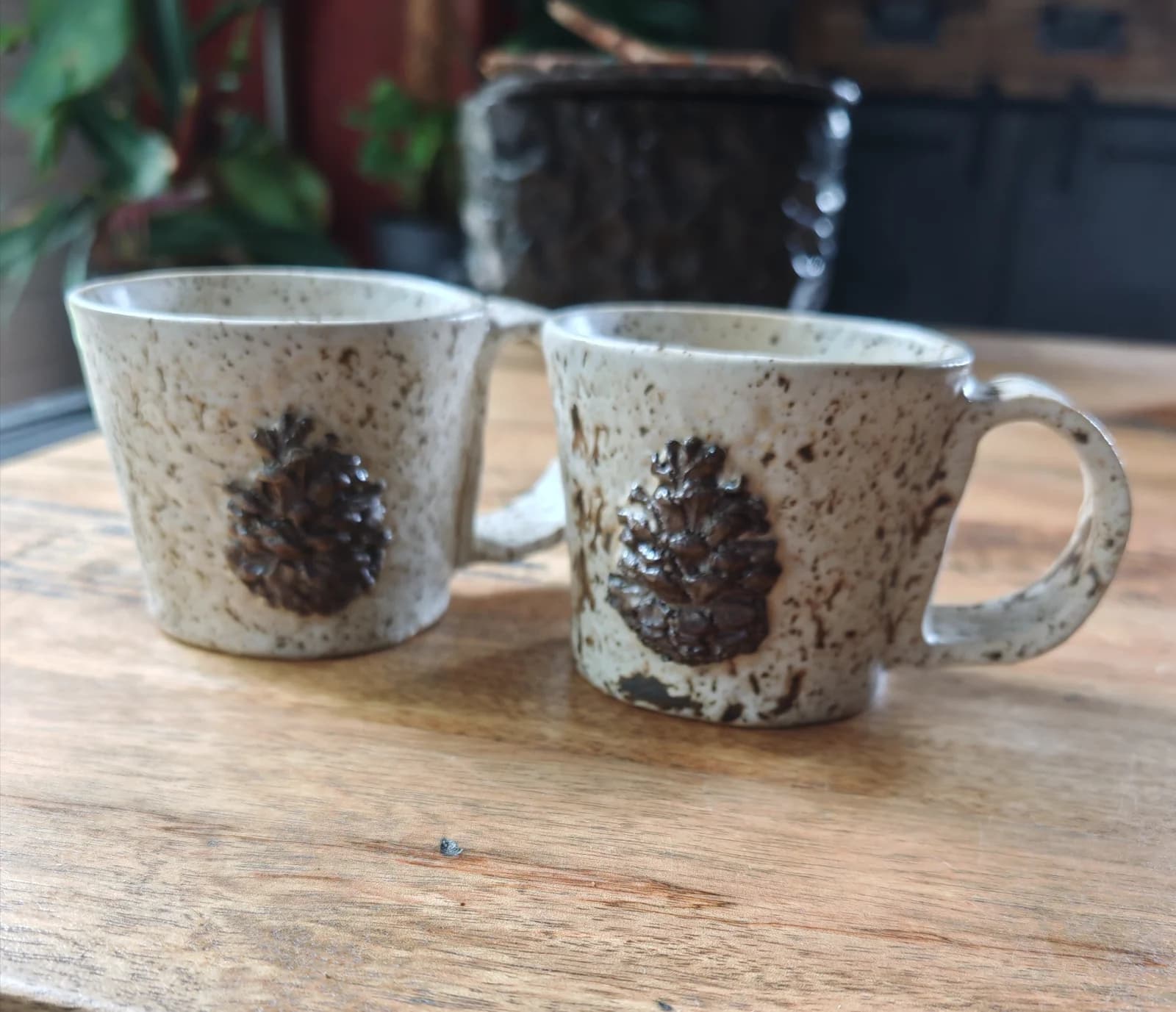 Deux tasses en céramique mouchetée avec motif pomme de pin en relief posées sur une table en bois.